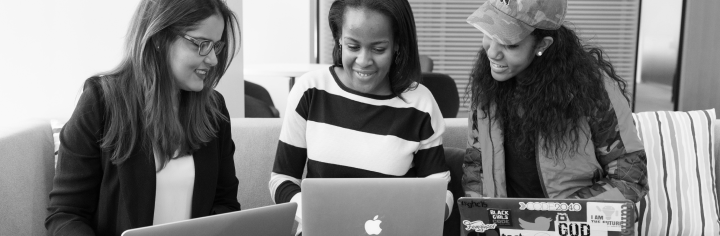 Three racially diverse women sitting on a sofa each with a
    laptop on their lap and smiling.