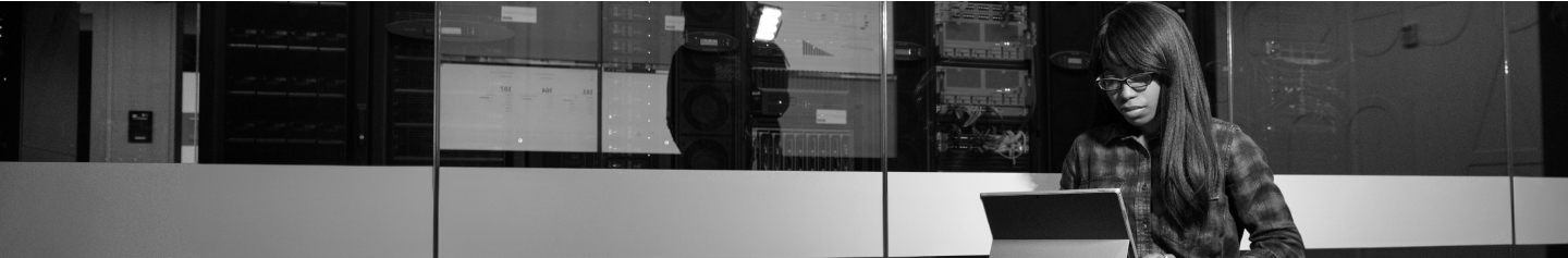 A black woman sitting in front of a server room while working on a laptop.